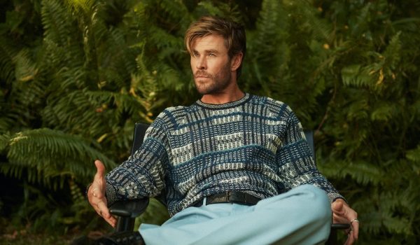 Chris Hemsworth usando uma blusa de manga comprida xadrez e calça de alfaiataria sentado em uma cadeira em frente a um fundo de plantas.